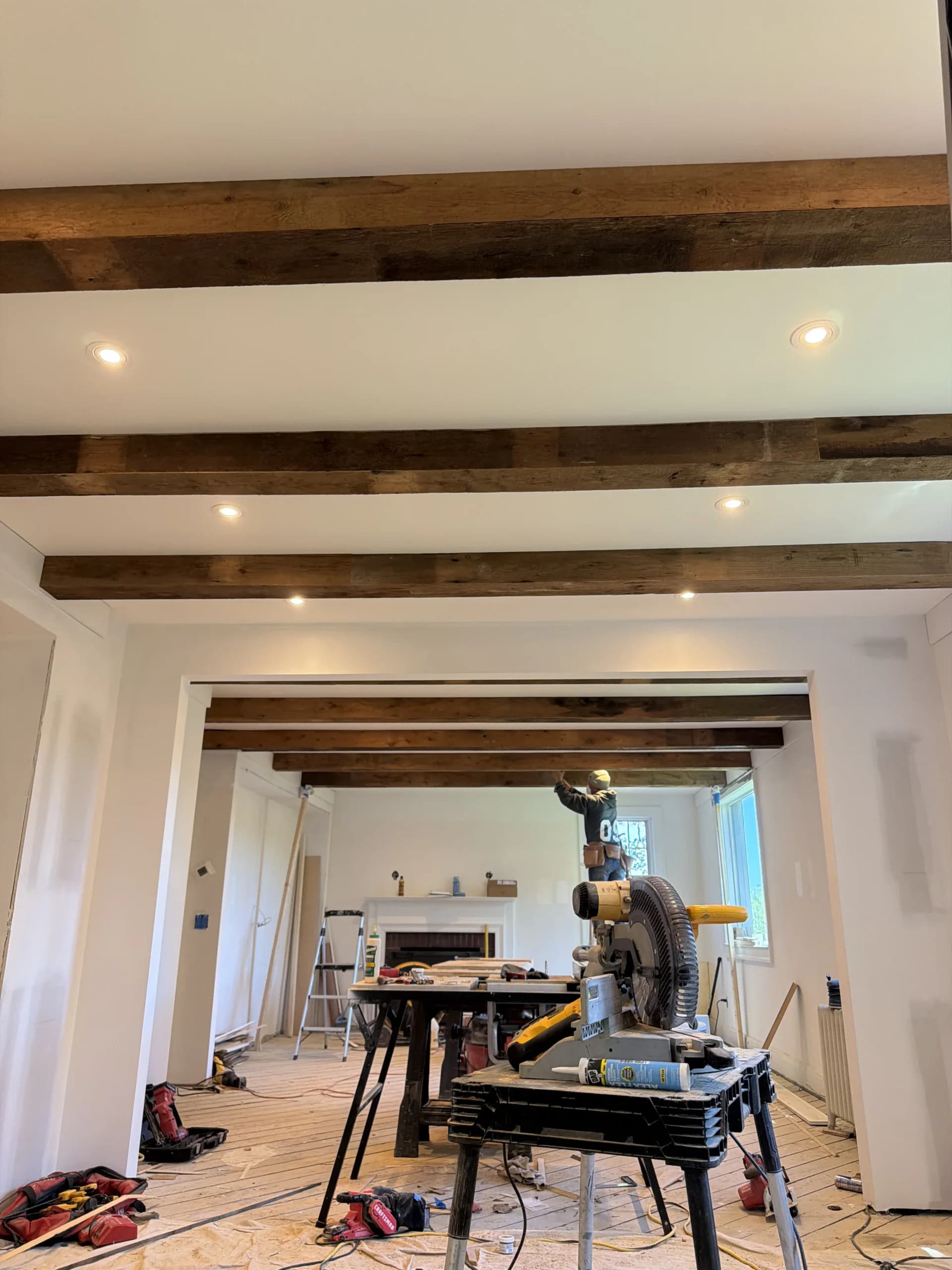 Open living space mid-renovation with exposed reclaimed wood ceiling beams and a miter saw on a stand - Greater Homes Solutions LLC