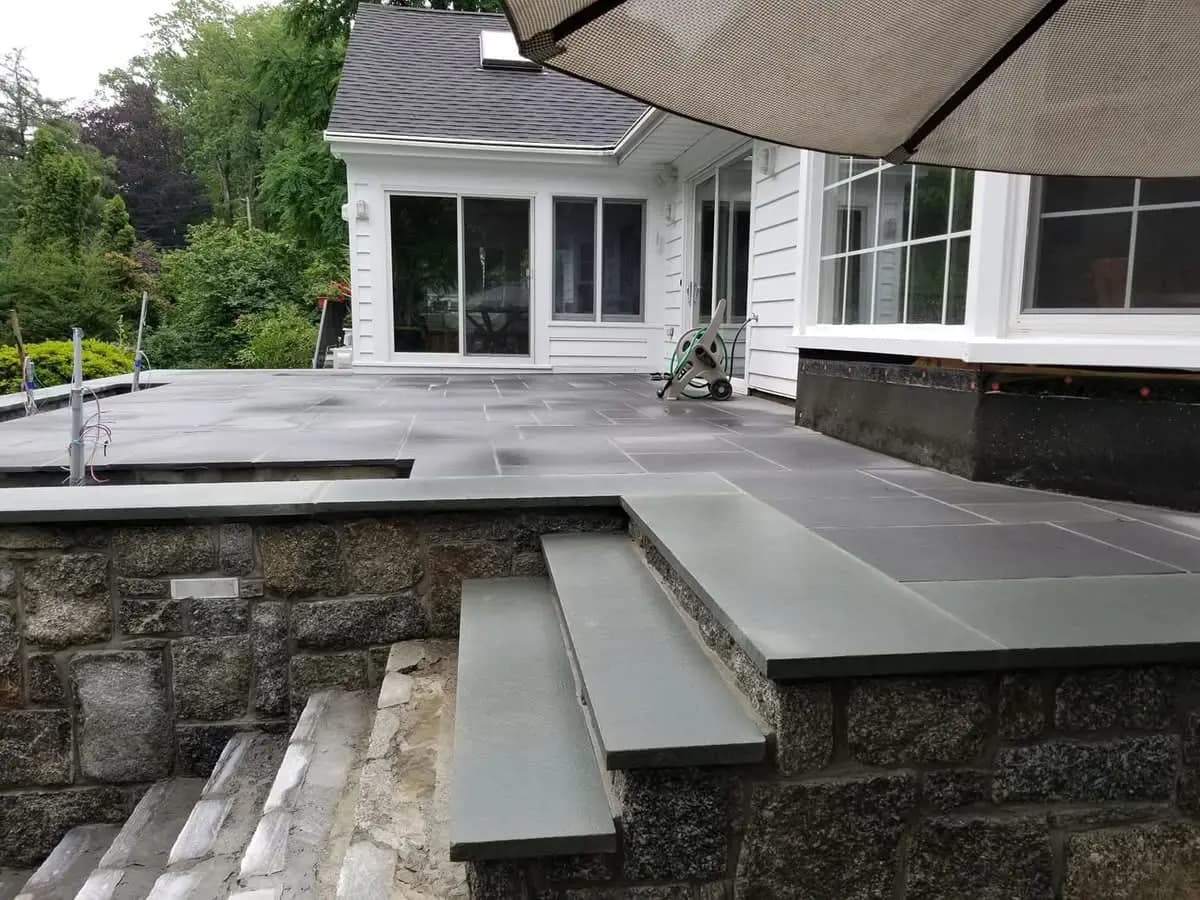 Bluestone patio and steps on a fieldstone retaining wall beside a white home - masonry by Greater Homes Solutions LLC