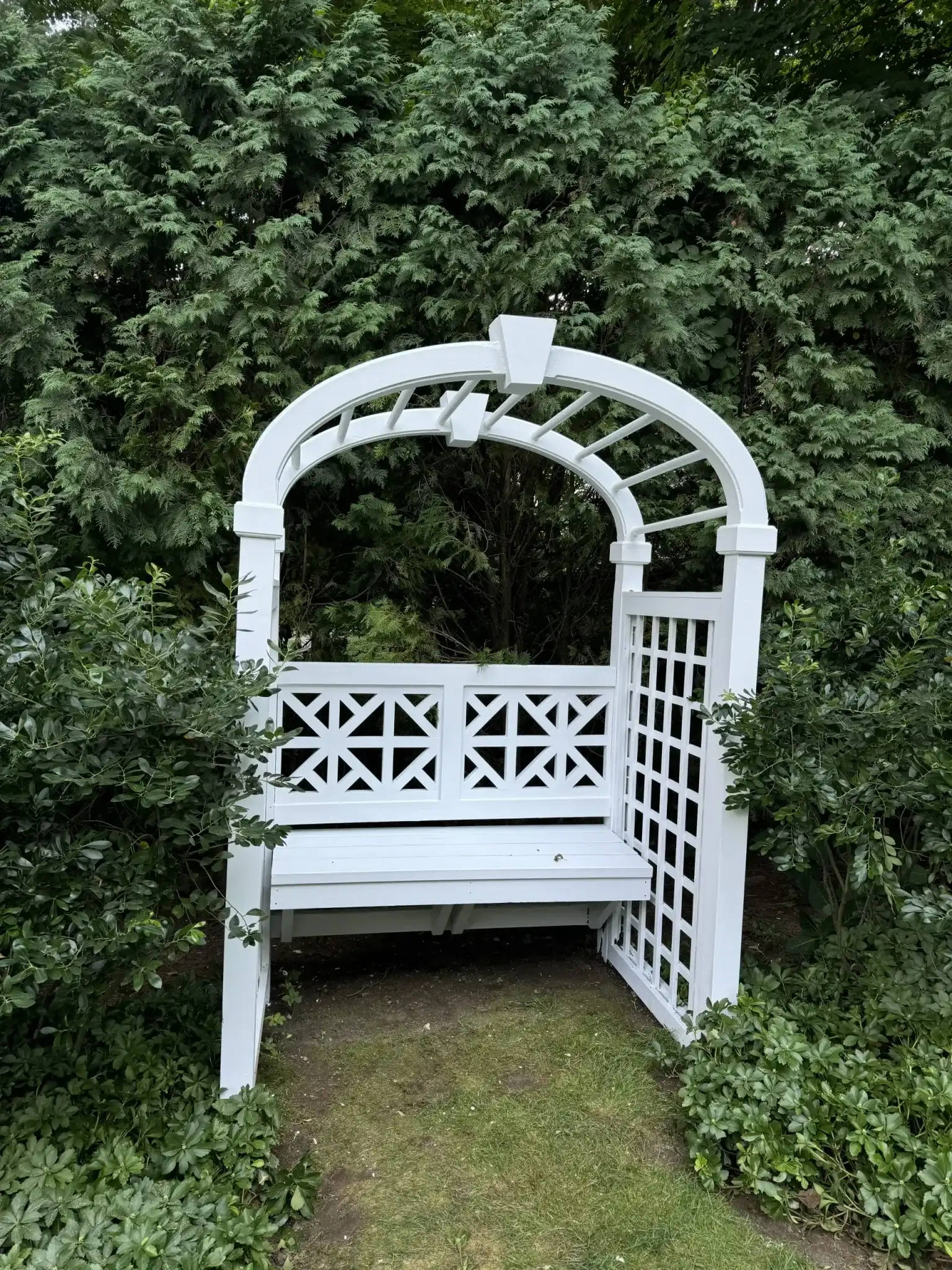 White garden arbor with a built-in bench set among evergreen hedges - Greater Homes Solutions LLC