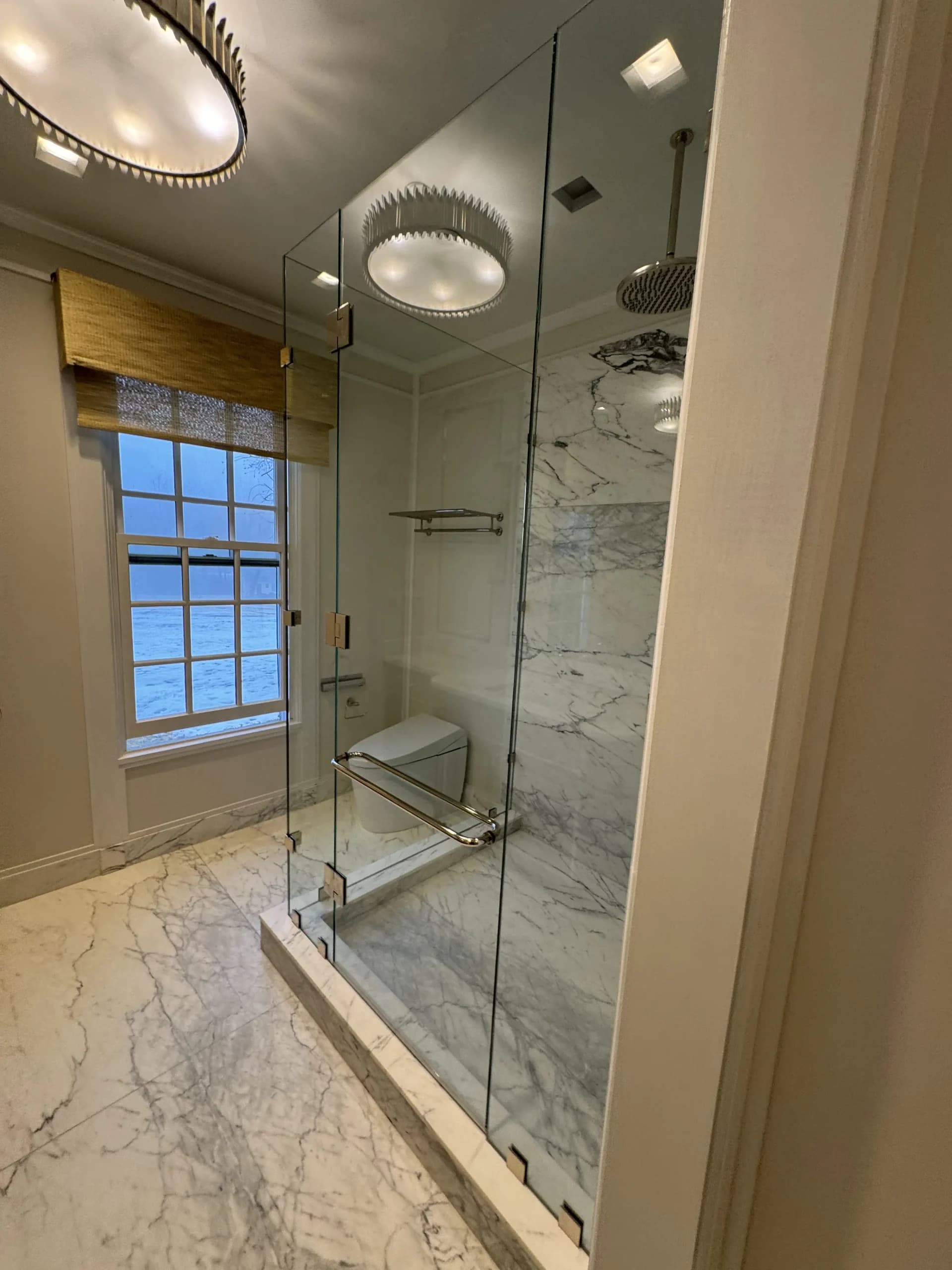 Marble-tiled shower with a rain showerhead behind a frameless glass door - bathroom remodeling by Greater Homes Solutions LLC