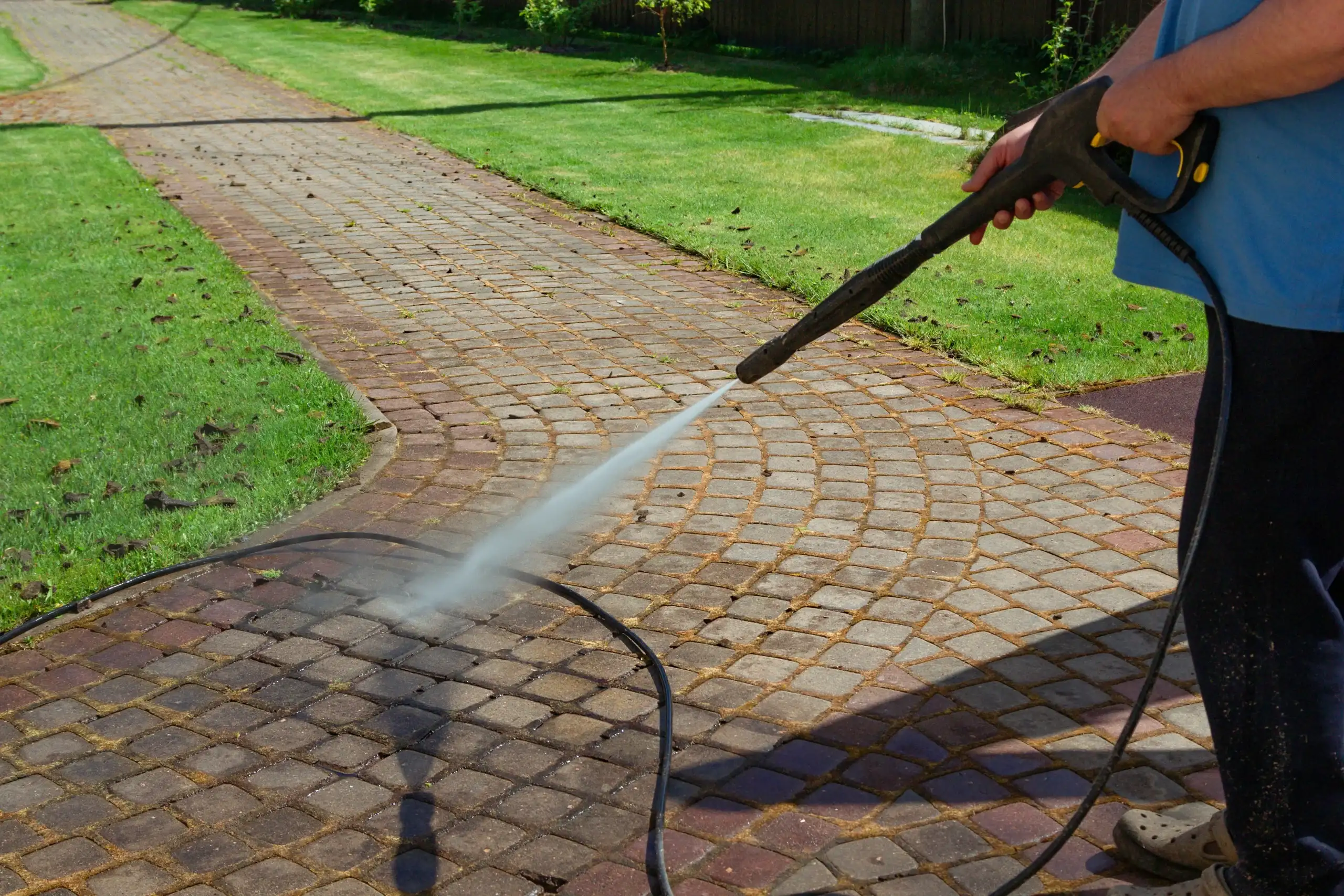 Pressure washer spraying a brick paver walkway beside a lawn
