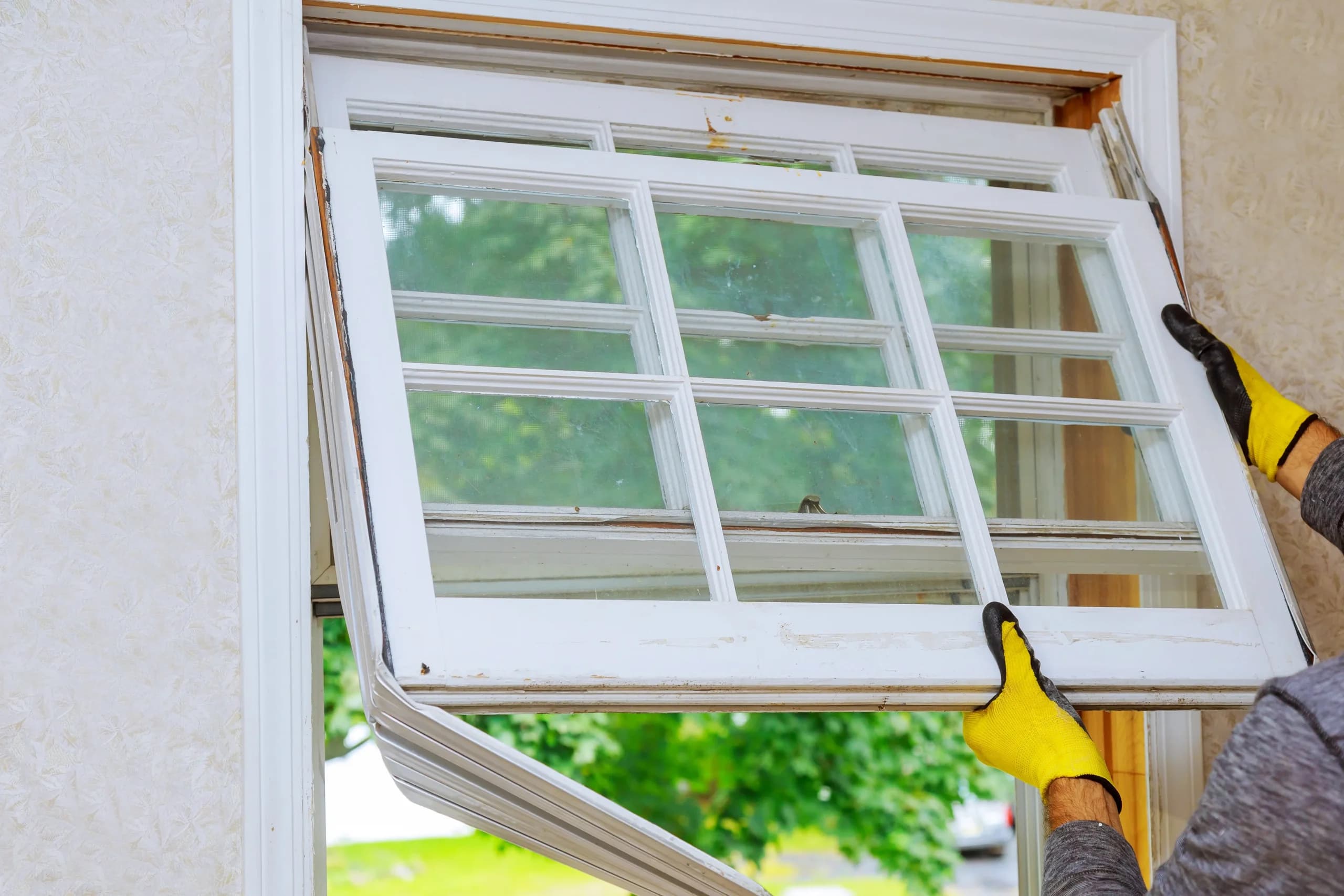 Installer in work gloves fitting a white double-hung window into a wall opening